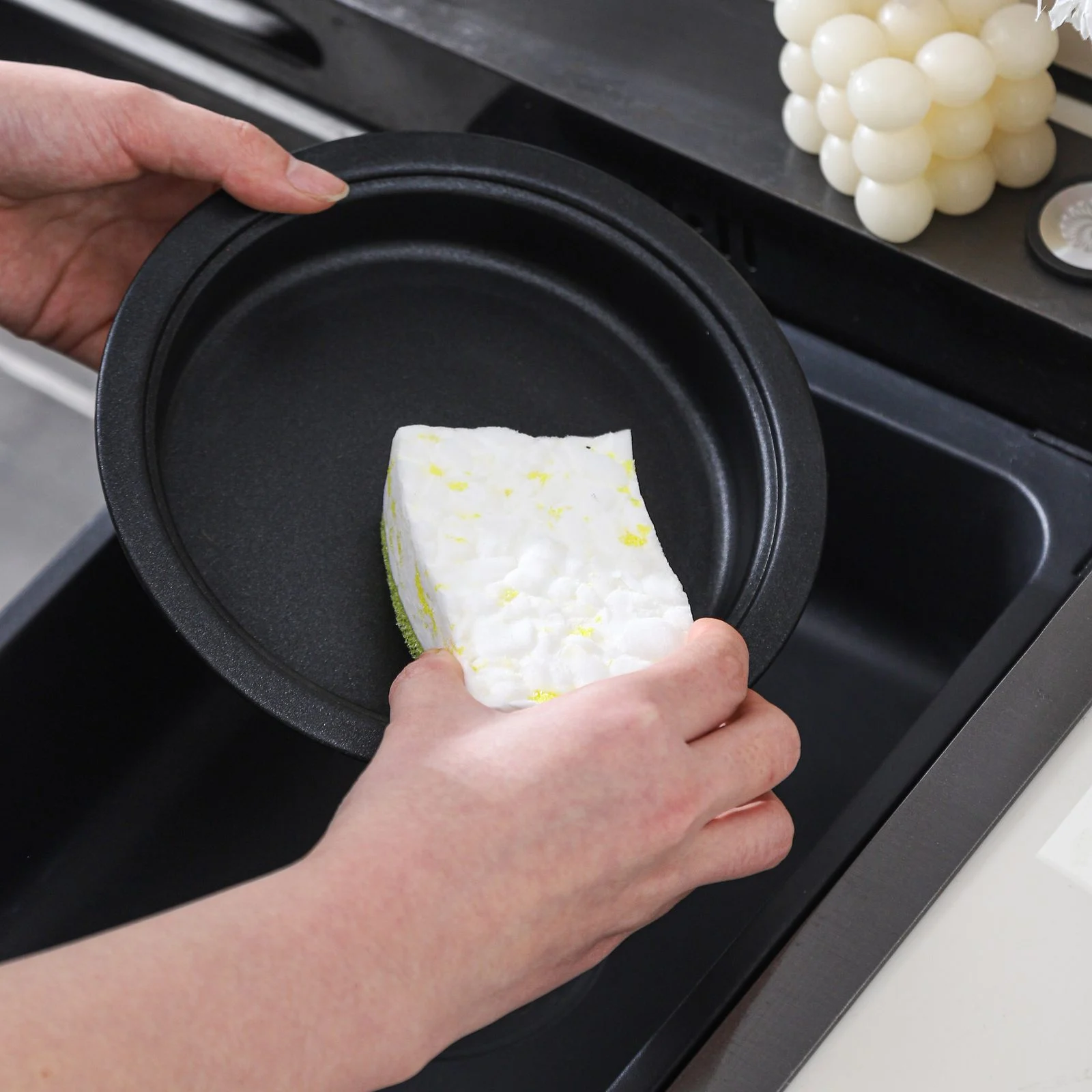 Hand scrubbing a black pan with a white green and yellow tri-layer sponge under running water